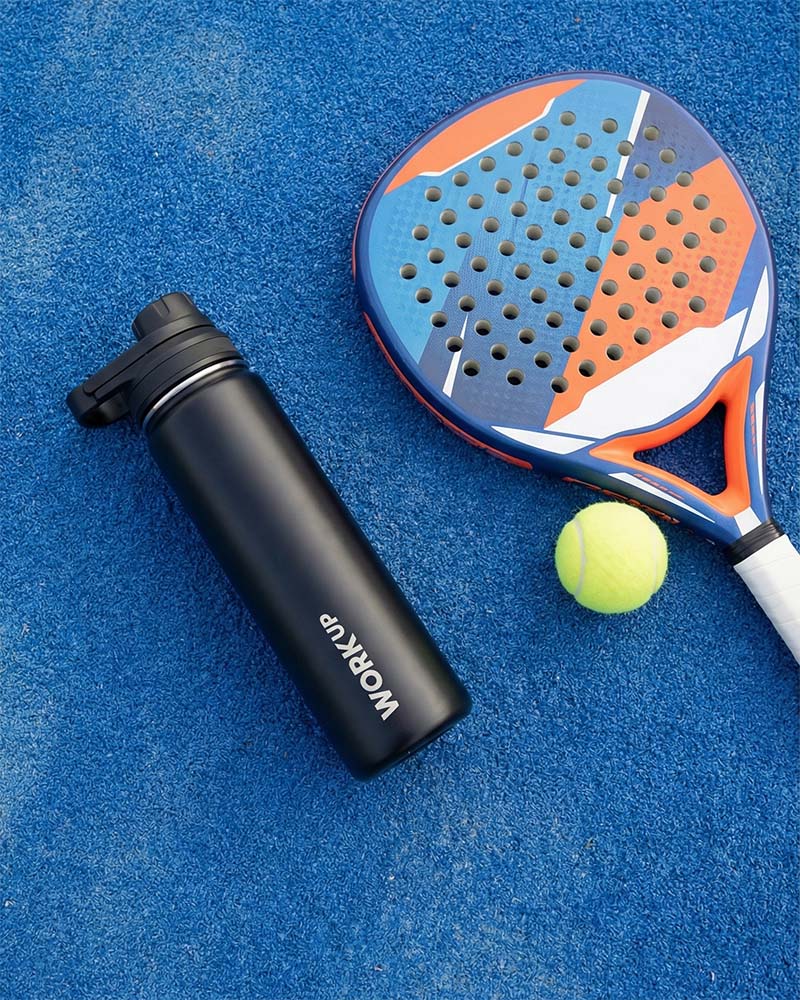 WORK UP-drikkeflaske ligger på jorden ved siden af en padelketcher og en bold. 