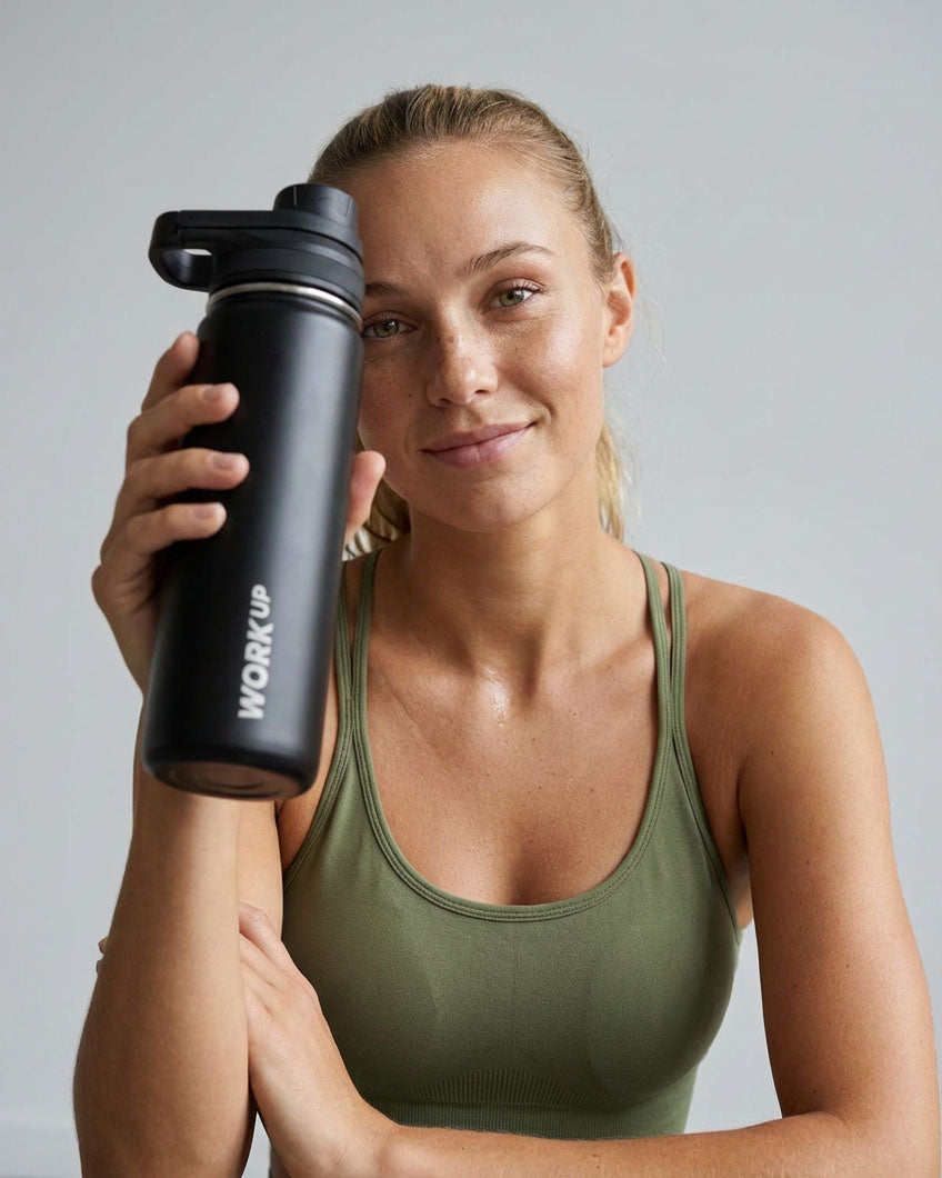 Kvinde i træningstøj holder en sort WORK UP-drikkeflaske i rustfrit stål og smiler mod kameraet.